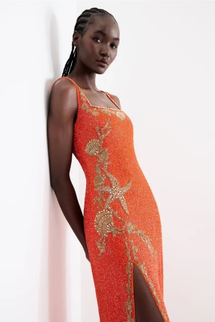 Model leaning on wall wearing tangerine orange maxi gown with gold coral beadwork