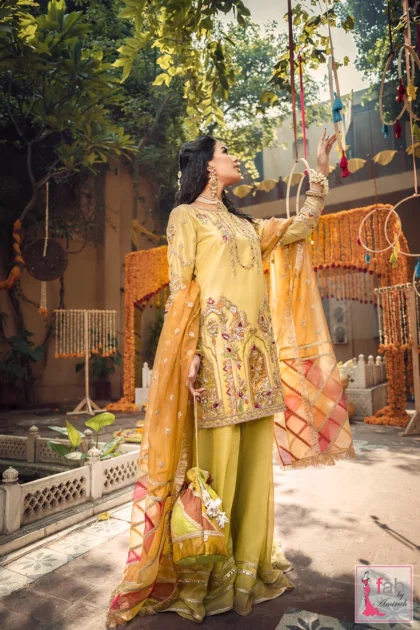 Model wearing a mustard Silk Mehndi Sharara Suit with an orange chatta patti dupatta and gold kiran fringe in a courtyard setting