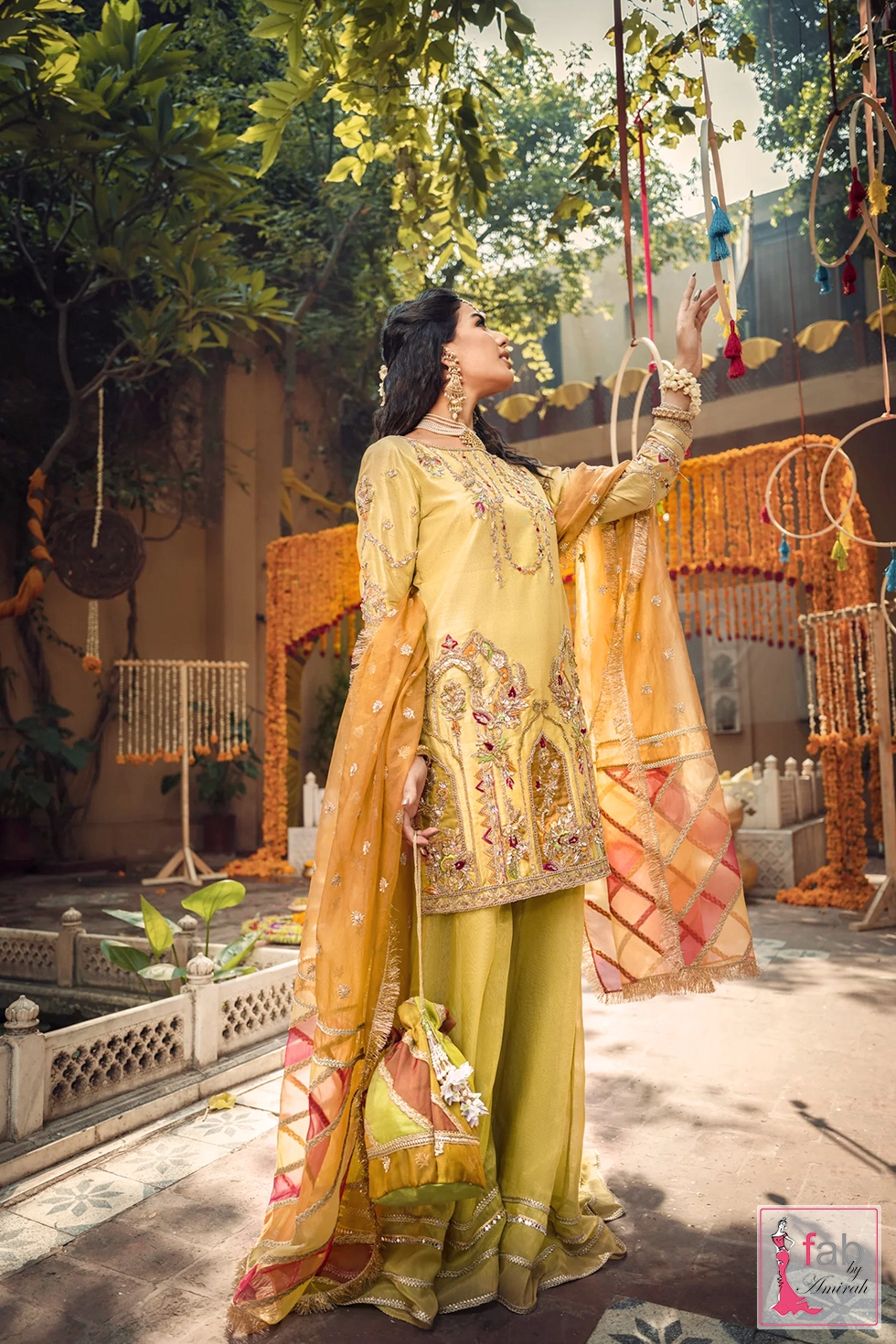 Model wearing a mustard Silk Mehndi Sharara Suit with an orange chatta patti dupatta and gold kiran fringe in a courtyard setting