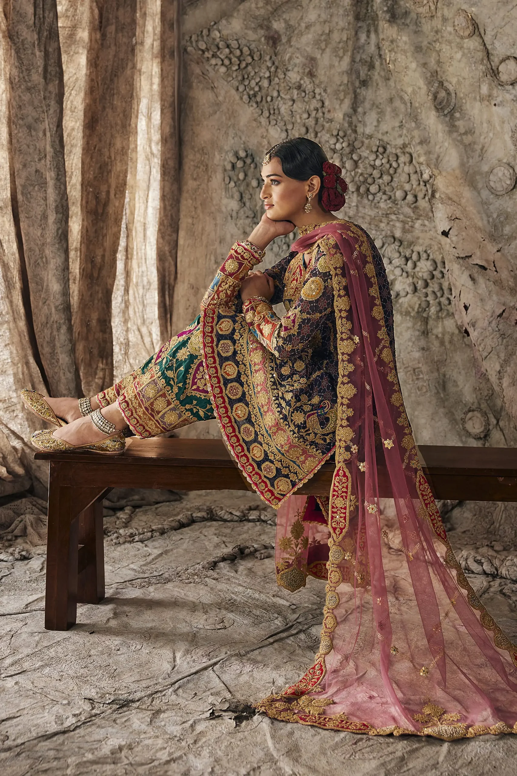 Side view of designer party wear dress showing model seated on bench with long flowing vintage rose net dupatta. Side view of designer party wear dress showing model seated on bench with long flowing vintage rose net dupatta.